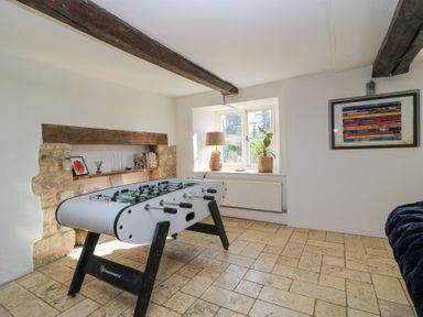 A room with a foosball table, a window with a lamp and plant, a built-in stone shelf with books and decor, a framed colorful artwork on the wall at Tickmorend Farm in Nailsworth