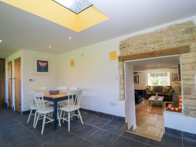 A dining area with a wooden table and six white chairs under a skylight next to a stone archway leading to a sitting room at Tickmorend Farm in Nailsworth