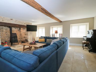 A living room with a blue sectional sofa a wooden coffee table a wood-burning stove a wall-mounted TV and a piano at Tickmorend Farm in Nailsworth