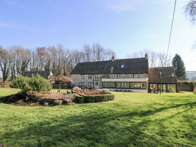 A stone house with multiple windows and green doors surrounded by grass and hedges at Tickmorend Farm in Nailsworth