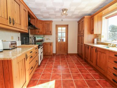 A kitchen with wooden cabinets red tiled floor white door and window at Buarthau Cottage in Llanrug