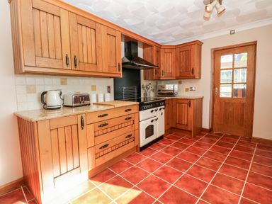 A kitchen with wooden cabinets a white oven a wooden door and red tiled floor at Buarthau Cottage in Llanrug