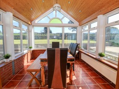 A dining area with wooden table and chairs in a conservatory with tiled floor and windows on all sides at Buarthau Cottage in Llanrug