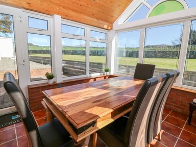A dining area with a wooden table and four black chairs in a sunroom with large windows at Buarthau Cottage in Llanrug