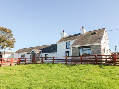 A house with white walls and a stone section with chimneys and a red wooden fence with grass in front at Buarthau Cottage in Llanrug