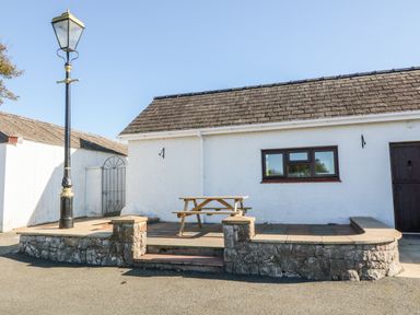A patio with a picnic table and stone wall next to a white building with a window and door at Buarthau Cottage in Llanrug