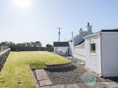 An exterior view of a white house with a lawn and stone wall at Buarthau Cottage in Llanrug