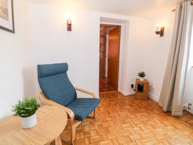 A room with a blue cushioned wooden chair a wooden side table with a plant a wooden speaker with a plant wooden parquet floor and a door at Buarthau Cottage in Llanrug