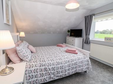 A bedroom with a bed covered in patterned bedding two bedside lamps a white dresser with a TV and a window with grey curtains at Buarthau Cottage in Llanrug