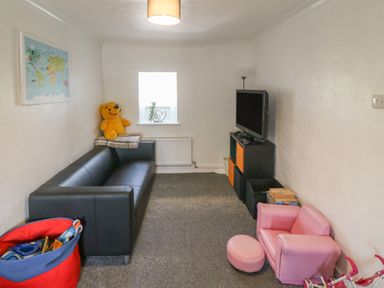 A living room with a black sofa a pink child's armchair and ottoman a television on a stand a window and toys at Buarthau Cottage in Llanrug