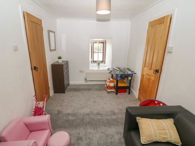 A playroom with two wooden doors a small window a chest of drawers a small foosball table toys a pink child sofa and a black armchair with a cushion at Buarthau Cottage in Llanrug
