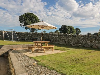 An outdoor garden area with a wooden picnic table and umbrella a stone wall and soccer goal in the background at Buarthau Cottage in Llanrug