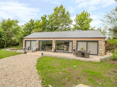 A single story stone house with large sliding glass doors and outdoor seating on a patio surrounded by trees at Coal Drops in Alnwick
