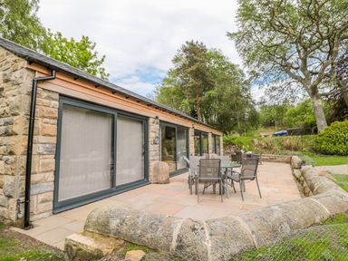 An outdoor patio with tables and chairs next to a stone building with large sliding glass doors surrounded by trees at Coal Drops in Alnwick