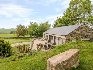 A stone building with large windows and outdoor seating on a patio surrounded by grass and trees at Coal Drops in Alnwick
