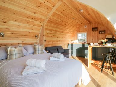 A wooden pod interior with a bed with pillows and towels a small sofa a kitchenette and a dining counter with stools at Chapel Pod in Callington