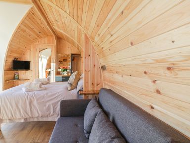 A wooden curved pod interior with a bed, a grey sofa, a wall-mounted TV, and a mirror with a dresser at Chapel Pod in Callington