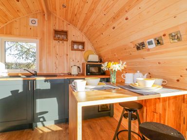 A small kitchen with wooden walls a window cabinets a microwave toaster kettle and a table set for two with dishes and flowers at Chapel Pod in Callington