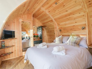A bedroom with a double bed towels on the bed a wall-mounted television and wooden curved walls at Chapel Pod in Callington