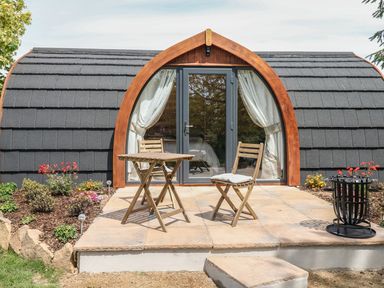An outdoor patio with a wooden table and two chairs in front of a small cabin with arched glass doors at Chapel Pod in Callington