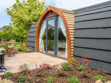 An outdoor patio area with wooden chairs and table next to a pod with arched glass doors and a garden bed with flowers at Chapel Pod in Callington