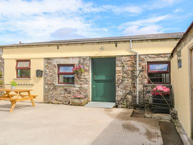 An outdoor area with a stone and yellow wall building with three windows and a green door a wooden picnic table and flower pots at The Old Cows House in Helston