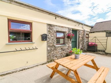 An outdoor patio area with a wooden picnic table and a potted plant a stone and plaster wall with windows a green door and flower baskets at The Old Cows House in Helston