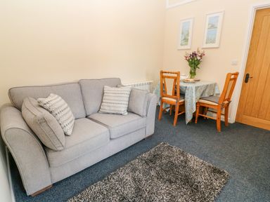 A living room with a grey sofa and pillows a small table with chairs flowers and paintings on the wall at The Old Cows House in Helston