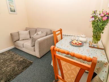 A small room with a gray sofa and a wooden table with two chairs a floral bouquet and tea set on the table at The Old Cows House in Helston