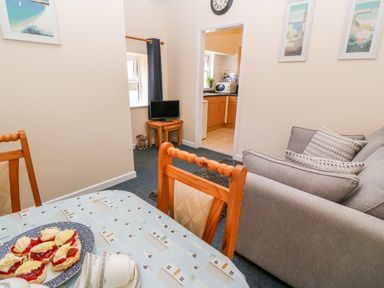 A dining table with scones and cups in a room with wooden chairs a gray sofa a small TV on nested tables and a view into a kitchen at The Old Cows House in Helston