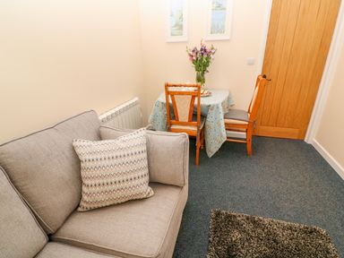 A small living room with a gray couch and a table with two wooden chairs and flowers at The Old Cows House in Helston