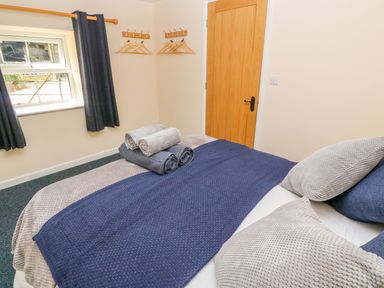 A bedroom with a bed covered in blue and beige blankets and pillows with rolled towels on top near a window with dark curtains and a wooden door at The Old Cows House in Helston