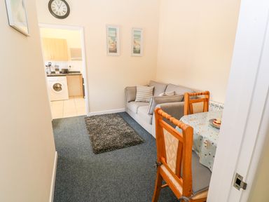 A small living room with a gray sofa pillows a round dining table with wooden chairs and a doorway leading to a kitchen at The Old Cows House in Helston