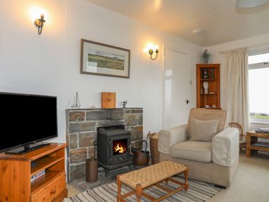 A living room with a stone fireplace and wood stove next to an armchair and wooden furniture at Allt y Mor in Harlech