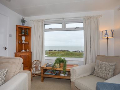 A living room with a large window showing a sea view two beige armchairs a wooden corner cabinet a wooden coffee table with a plant and magazines and a floor lamp with a lobster design shade at Allt y Mor in Harlech