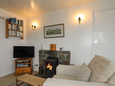 A living room with a beige armchair a wooden TV stand with a TV a stone fireplace with a lit stove and wall-mounted bookshelf at Allt y Mor in Harlech