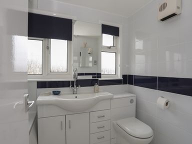 A bathroom with a white sink and cabinet a toilet and two windows with dark blinds at Allt y Mor in Harlech