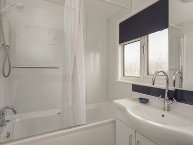 A bathroom with a bathtub shower combination a white shower curtain a window with a black blind and a white sink with a chrome faucet at Allt y Mor in Harlech