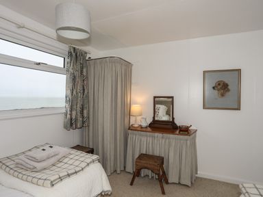 A bedroom with a window showing a sea view a bed with folded towels a dressing table with a mirror lamp and a wooden stool a framed dog painting on the wall at Allt y Mor in Harlech