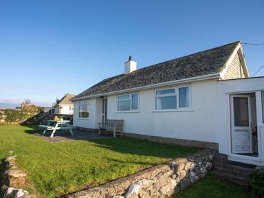 A white house with a stone roof in a garden with a picnic table and wooden bench at Allt y Mor in Harlech