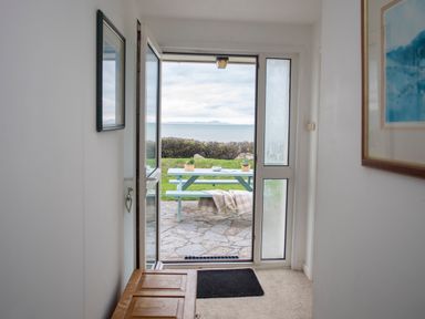 An open door leading to a stone patio with a blue picnic table overlooking a grassy area and the sea at Allt y Mor in Harlech