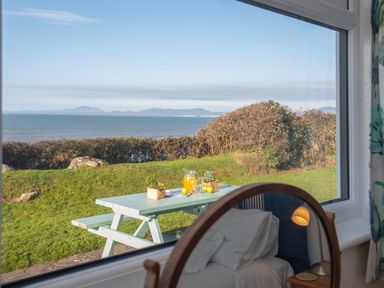 A window view of a picnic table with a jug of orange juice and apples on green grass by the sea at Allt y Mor in Harlech