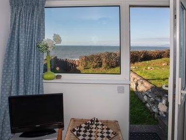 A window with a vase of flowers on the sill a television and a chessboard on a table with a view of the sea and a garden at Allt y Mor in Harlech