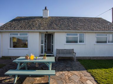 A white house with a door and windows a wooden bench and a picnic table with a jug of orange juice on a stone patio at Allt y Mor in Harlech