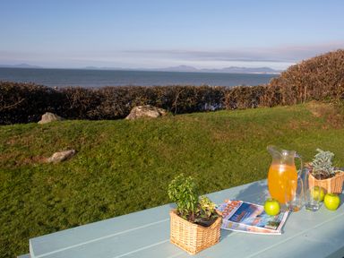 A picnic table with a jug of orange juice apples glasses magazines and potted plants facing the sea at Allt y Mor in Harlech