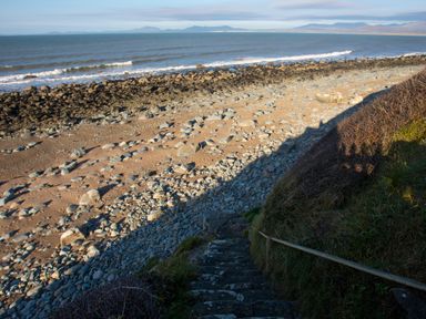 Stone steps leading down to a rocky beach with waves and mountains in the distance at Allt y Mor in Harlech