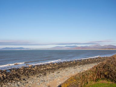 A rocky beach with waves and distant hills under a clear blue sky at Allt y Mor in Harlech