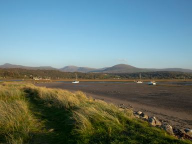A grassy path leading to a beach with boats and mountains in the background at Allt y Mor in Harlech