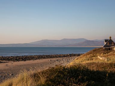 A rocky beach with a house on a grassy hill and mountains in the background at Allt y Mor in Harlech