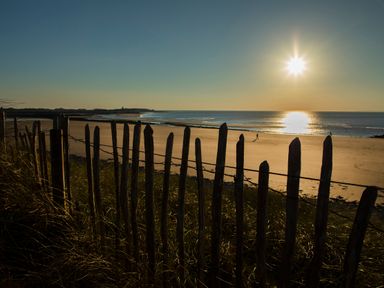 A wooden fence next to a sandy beach with the sun low in the sky and a person walking near the shoreline at Allt y Mor in Harlech